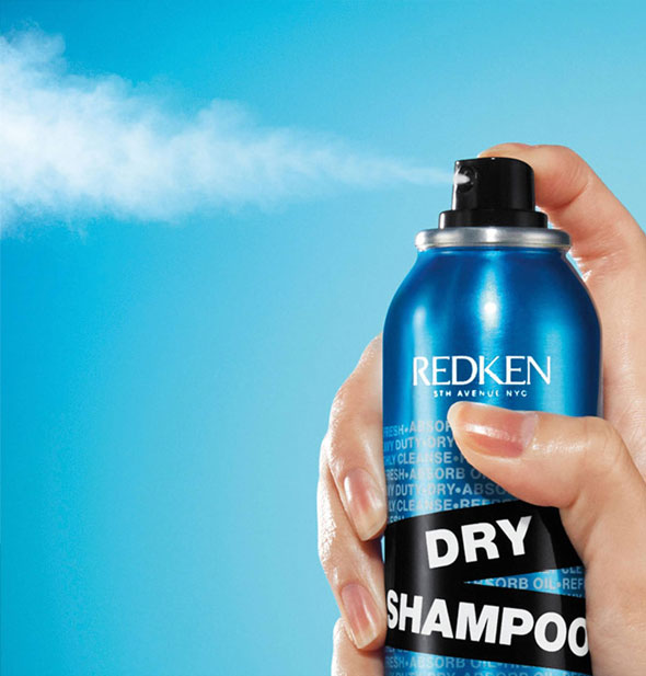 Model's hand dispenses a fine plume of Redken Dry Shampoo against a blue background