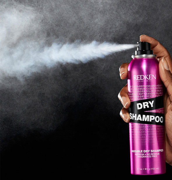 Model's hand dispenses a fine powdery plume from a can of Redken Invisible Dry Shampoo against a black background