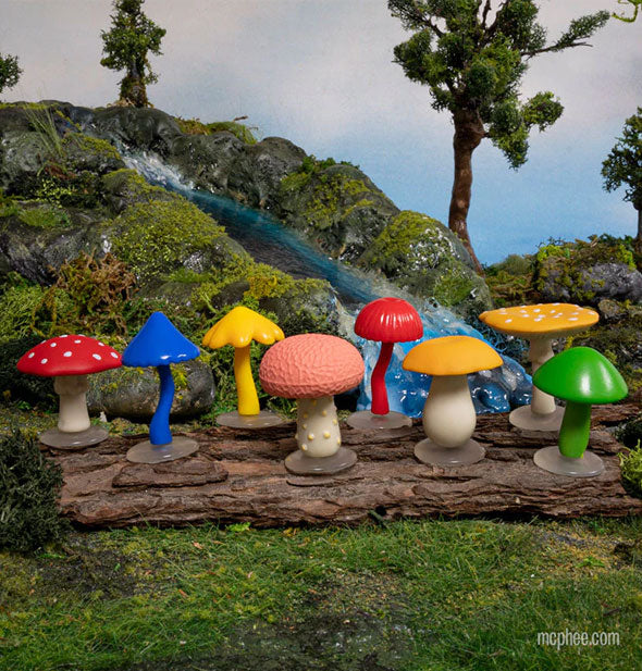 Mushroom figurines on a log staged against a mossy backdrop with babbling brook