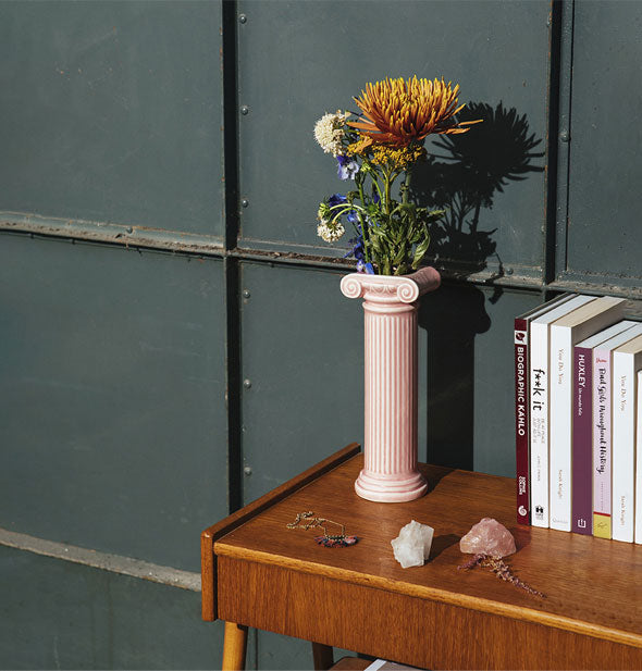 Pink column vase on a wooden desktop staged with books and crystals holds a small bouquet of wildflowers against a dark green industrial-looking wall