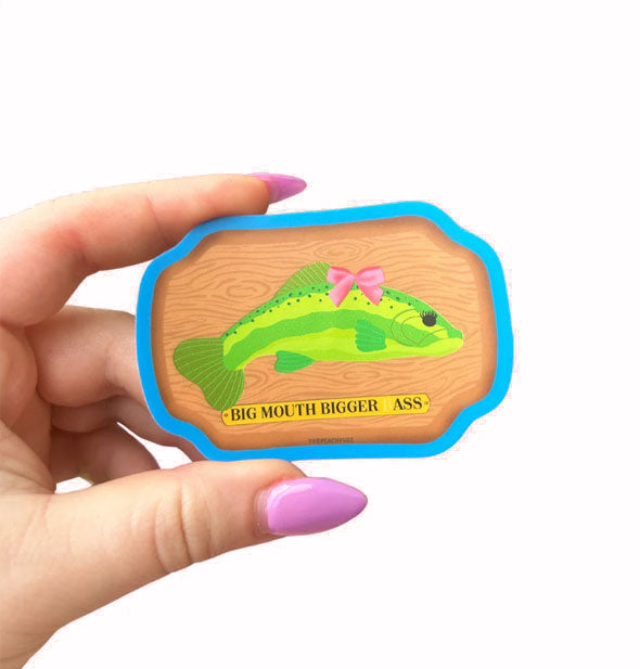 Model's hand holds a sticker designed as a Big Mouth Billy fish plaque but the fish is wearing a pink bow and the caption says, "Big Mouth Bigger Ass"