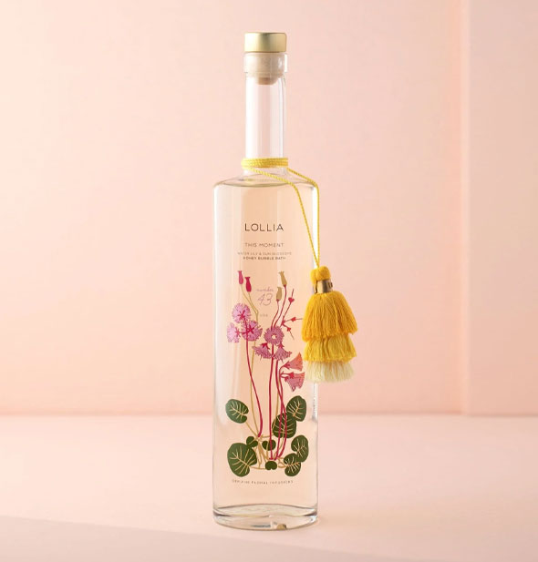 Long, slender, clear glass bottle of Lollia This Moment Bubble Bath with floral design and tiered yellow tassels hanging from the neck