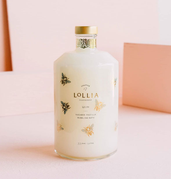 Wide cylindrical glass bottle of Lollia Wish Sugared Pastille Bubbling Bath is filled with milky-white contents and features metallic gold bee accents