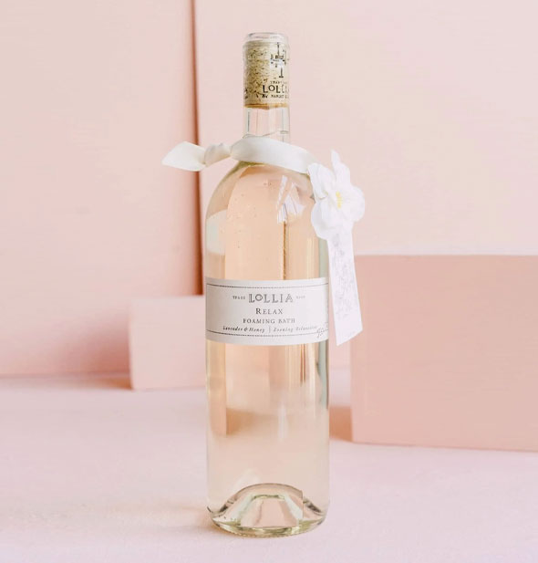 Clear Lollia Relax Foaming Bath glass bottle is topped with a cork and decorated with a white ribbon and flower sash around its neck