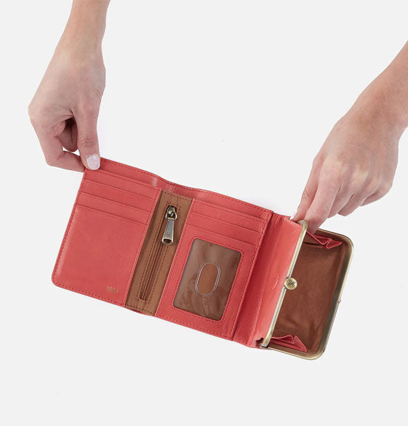 Model's hands hold open a salmon leather trifold wallet with brass hardware and brown lining