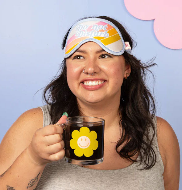 Smiling model holding a glass daisy coffee cup wears a Do Not Disturb sleep mask up above eyes covering forehead
