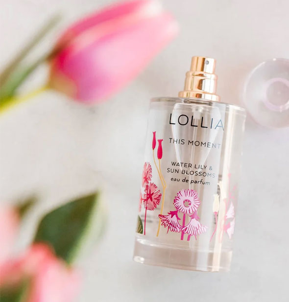 Lolia This Moment Eau de Parfum bottle laying on its side with cap removed and a pink tulip with greenery blurred in the foreground