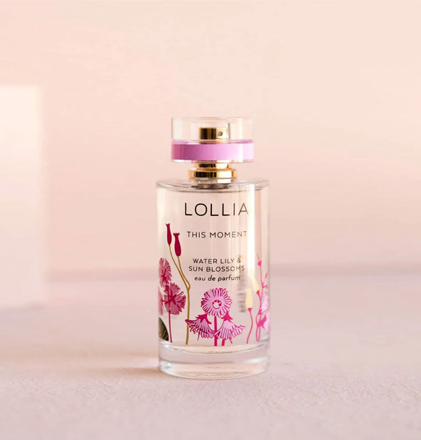 Cylindrical clear glass Lollia: This Moment Water Lily & Sun Blossoms Eau de Parfum bottle with pink floral motif and pink accent stripe on its clear glass lid that covers a gold nozzle