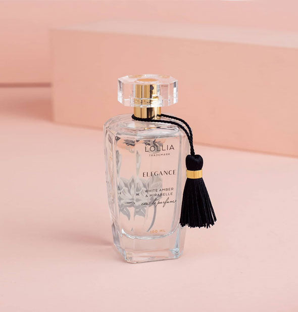 Clear bottle of Lollia Elegance Eau de Parfum on a pink backdrop features a faceted lid over gold spritzer, subtle black and white floral patterning, and black tassel hanging from the neck