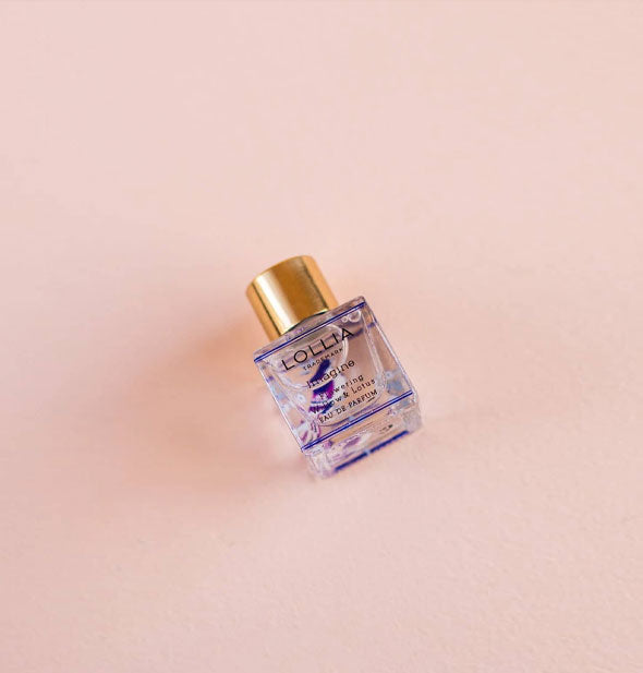 Small square glass Lollia Imagine Eau de Parfum bottle with blueish-purple design details and gold cap rests on a pink surface