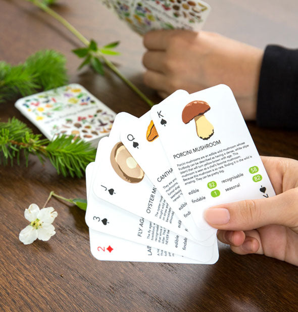 Models hold sample spreads from the Foragers Playing Cards deck at a wooden tabletop scattered with flowers and branches