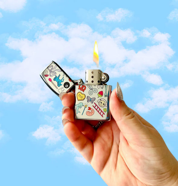 Model's hand holds a lit silvery flip-top lighter printed with playful graphics up in front of a blue sky with clouds backdrop