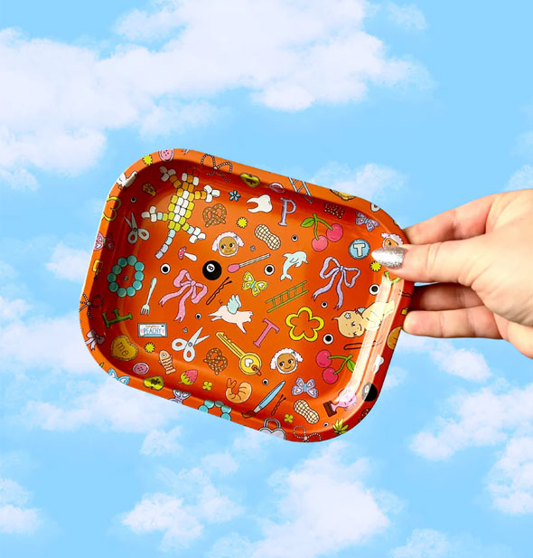 Model's hand holds a rectangular orange tray with rounded corners and illustrations of random objects all over it in front of a backdrop of blue sky with clouds