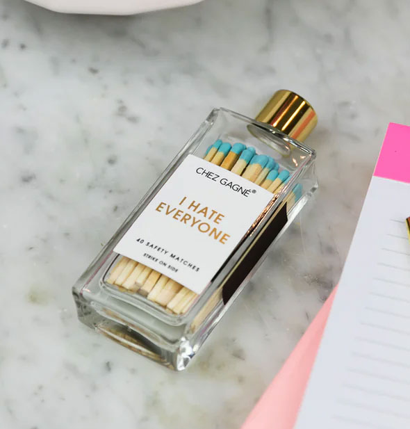 I Hate Everyone safety match bottle lays on its side on a marble surface next to a lined notepad
