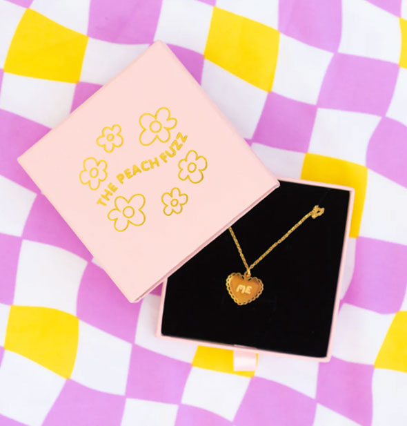 Gold necklace with heart-shaped pendant that says "ME" inside a pink The Peach Fuzz gift box on a wavy purple, white, and yellow checker print backdrop