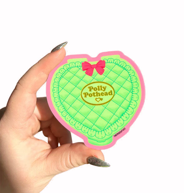 Model's hand holds a heart-shaped sticker designed to resemble a green Polly Pocket toy compact with pink bow and metallic gold label in the center that says, "Polly Pothead"