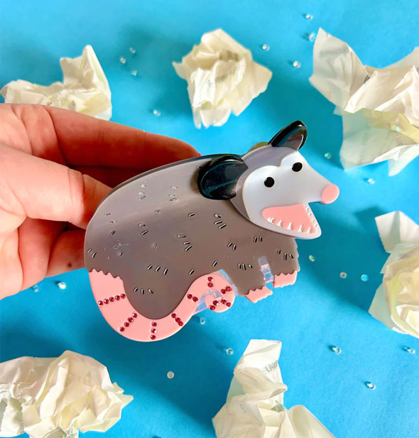 Model's handholds a hair clip designed to resemble an opossum with mouth open and small red rhinestone details on its tail; balled up paper is scattered on a blue surface in the background