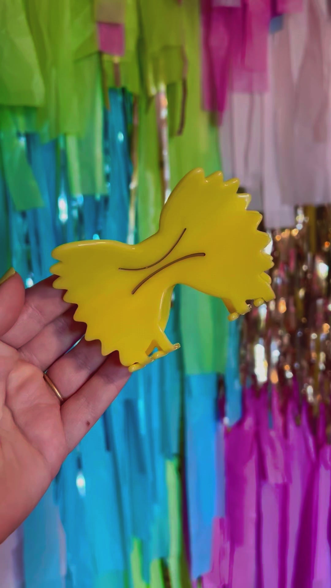 Video shows model's hand opening and closing a farfalle pasta hair clip against a multicolored tinsel backdrop