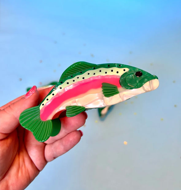 Model's hand holds a pink, white, and green rainbow trout hair clip with black rhinestone spot accents