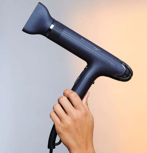 Model's hand holds a Bio Ionic Smart-X hair dryer against a pastel blue-to-orange ombre backdrop