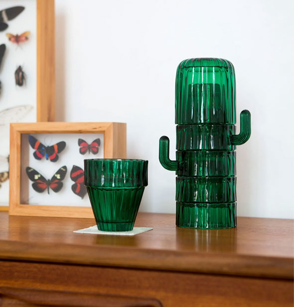 Stackable green saguaro cactus drinking glasses on a wooden tabletop next to mounted butterfly shadow boxes