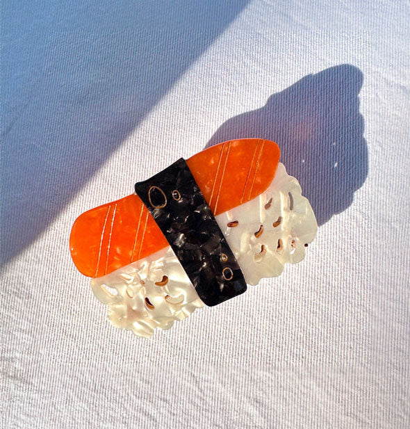 White, black, and orange clip with gold line details designed to resemble a sushi roll rests on a light denim surface
