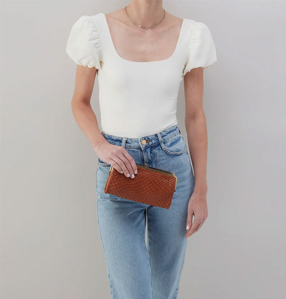 Model in jeans and a white blouse stands holding a brown woven leather wallet with gold-toned frame hardware