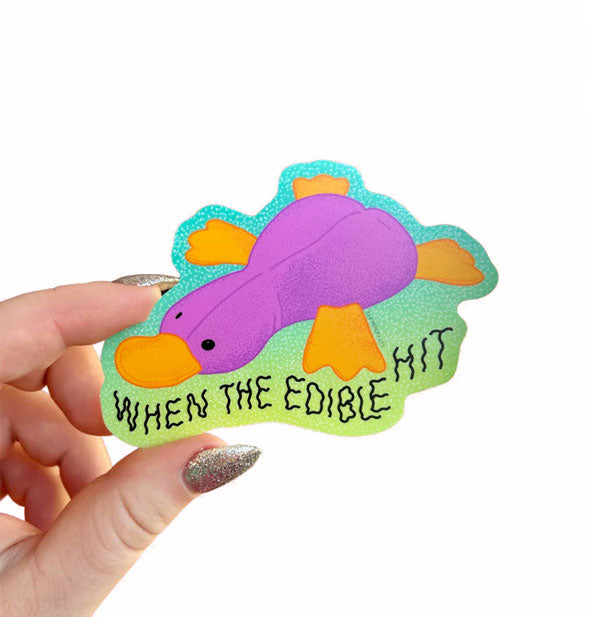 Model's hand holds a green ombre sticker with purple and orange platypus illustration that says, "When the edible hit" in wavy black lettering
