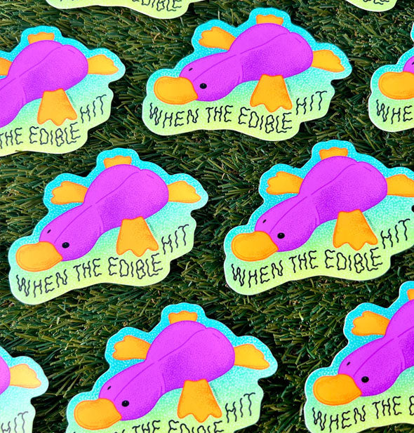 Several When the Edible Hit platypus stickers rest on green astroturf