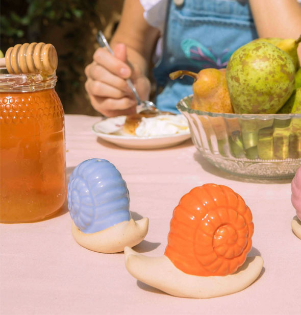 Blue- and orange-shelled ceramic snail salt and pepper shakers rest on a pink cloth-covered outdoor table set with honey and fresh fruit; a model with dessert plate is in the background