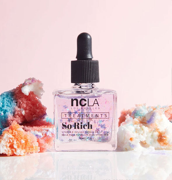 Square glass bottle of NCLA Treatments So Rich cuticle oil with black rubber dropper cap is staged with pieces of rainbow cake