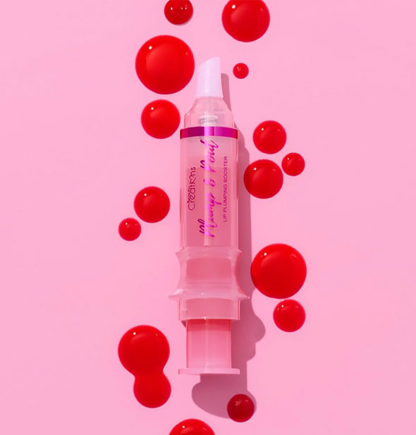 A syringe-shaped tube of Beauty Creations Plump & Pout lip gloss surrounded by bright orange-red droplets of product