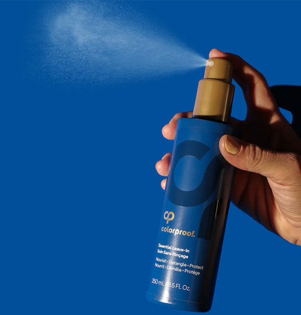 Model's hand dispenses a fine mist of ColorProof Essential Leave-In into the air against a blue background