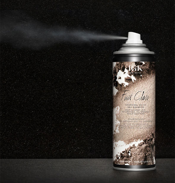 An application of IGK First Class Charcoal Detox Dry Shampoo is dispensed from can against black background