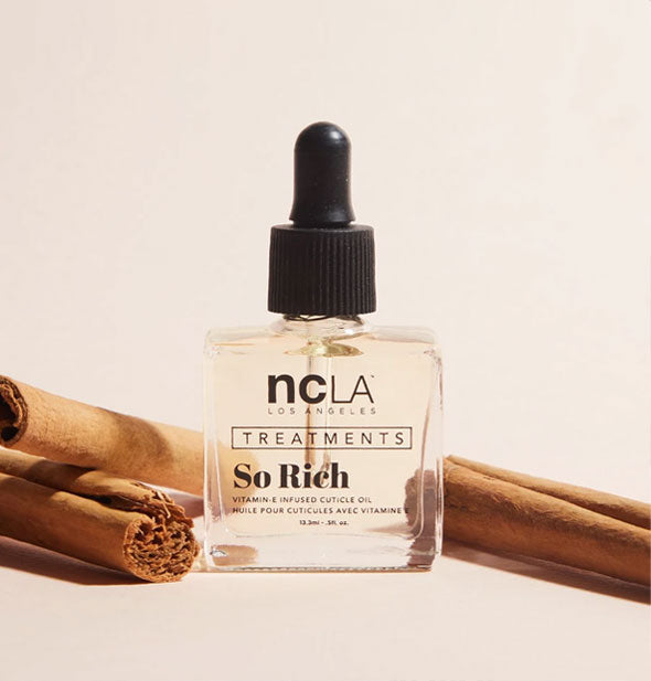 Square glass bottle of NCLA Treatments So Rich cuticle oil with black rubber dropper cap is staged with sticks of cinnamon