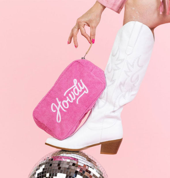 Model wearing a white cowgirl boot propped on a disco ball holds the pink Howdy pouch by its zipper tab