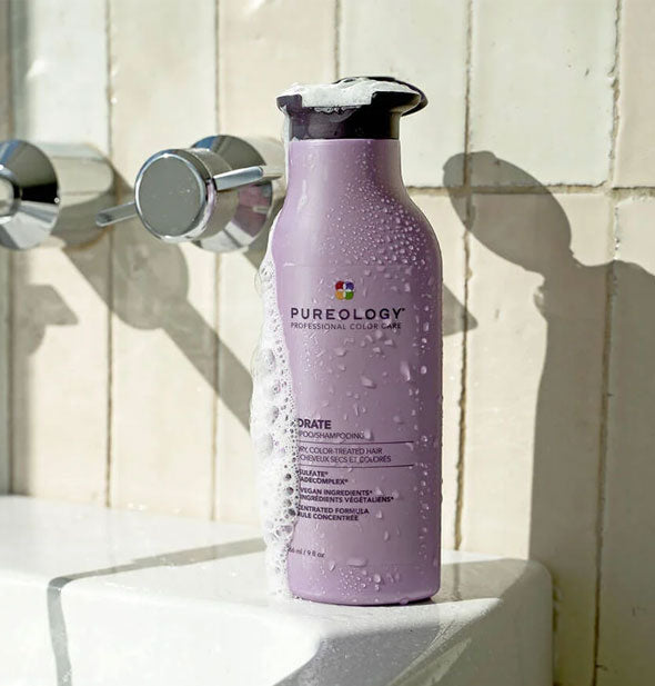 A bottle of Pureology Hydrate Shampoo with lather running down the side sits on a bathtub ledge