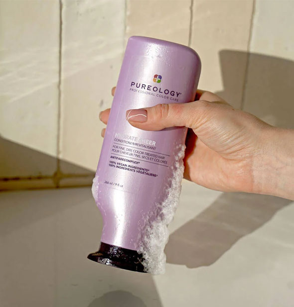 Model's hand holds a bottle of Pureology Hydrate Sheer Conditioner with some lather running down the side