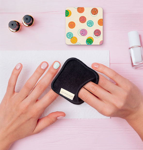 Model demonstrates use of a reusable nail polish remover pad on a surface staged with nail polish bottles and a square smiley face print emery board