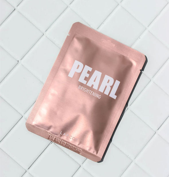 Rosy colored Pearl Brightening face mask foil pack rests on a white tile surface