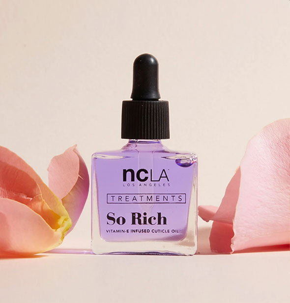 Square glass bottle of NCLA Treatments So Rich cuticle oil with black rubber dropper cap is staged with pink rose petals