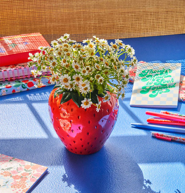 Strawberry flower vase is filled with small white daisies and is staged with colorful notebooks and pens on a blue surface in front of a brown window blind
