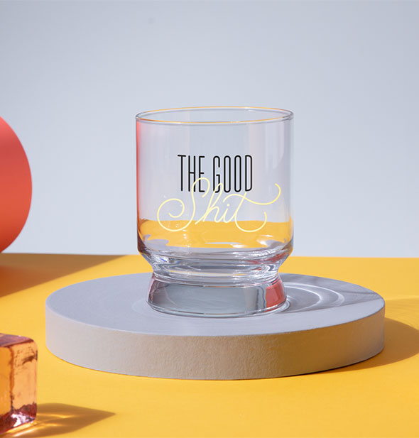 The Good Shit rocks glass sits atop a round pedestal on a colorful background