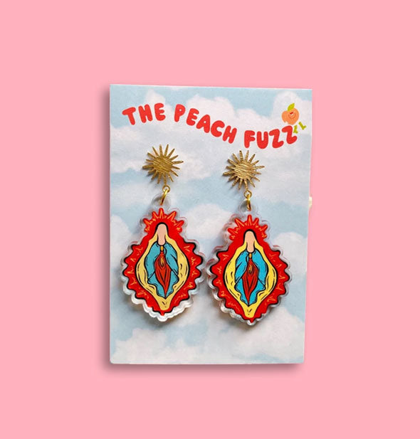 Acrylic earrings feature artwork that resembles vulvae dangling from gold sun-shaped posts on blister card printed with, "The Peach Fuzz"