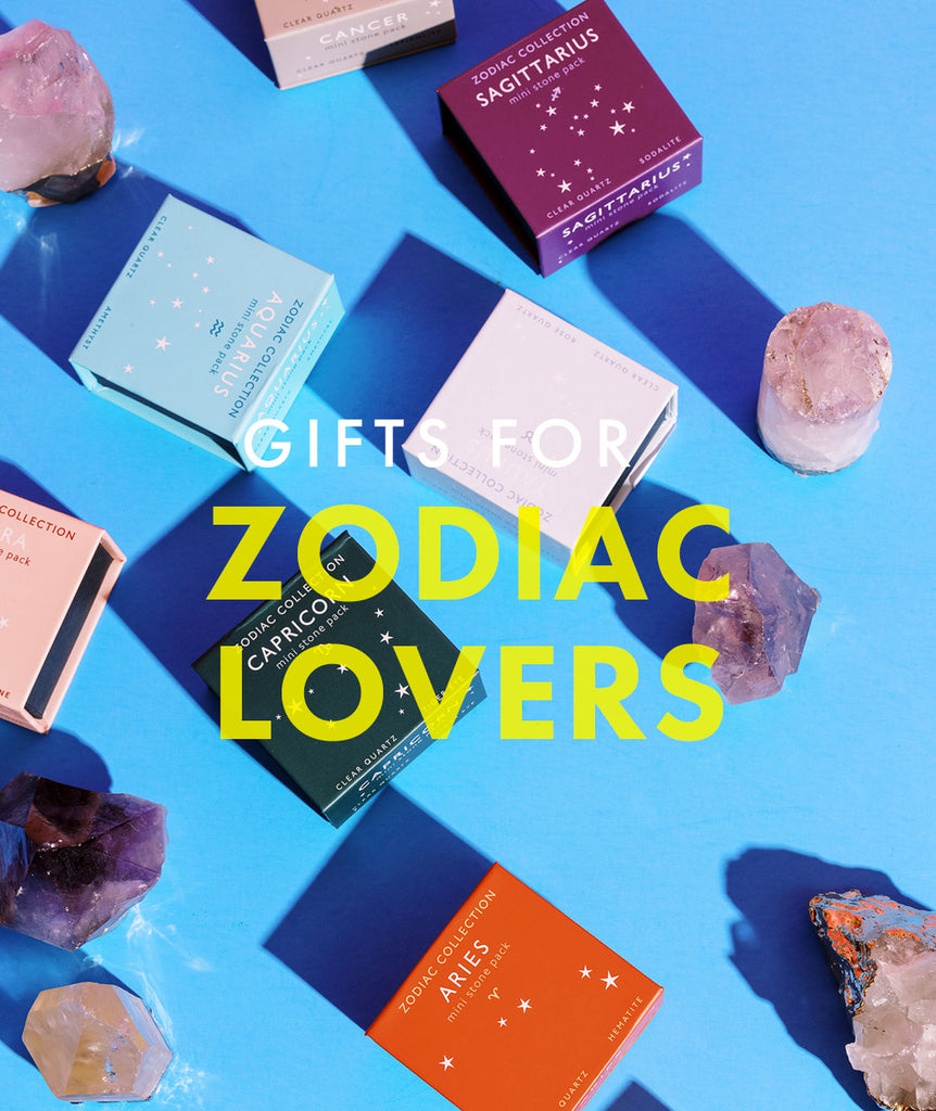 Crystals and boxes printed with astrological signs on a blue surface back the words, "Gifts for Zodiac Lovers" in white and yellow lettering