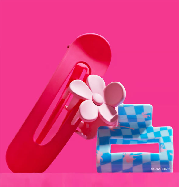Three hair clips against an intense pink background: one dark pink elongated oval shape, one light pink flower shaped, and one rectangular blue checker print