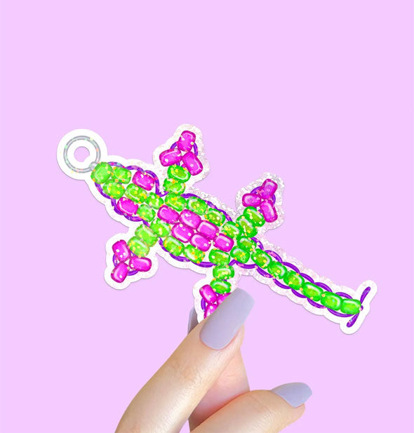 Model's hand holds a sticker designed as a pink and green beaded lizard keychain reminiscent of the 1990s