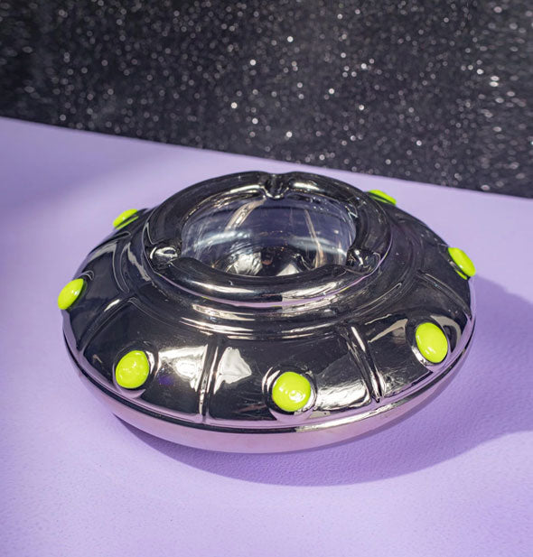 Shiny black UFO-shaped ashtray with green glow-in-the-dark circle accents rests on a purple surface against a dark speckled backdrop