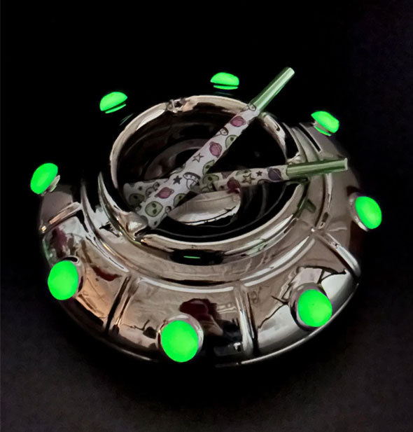 Shiny black UFO-shaped ashtray with green glow-in-the-dark circle accents rests on a black surface and holds two rolled cigarettes with space-themed paper