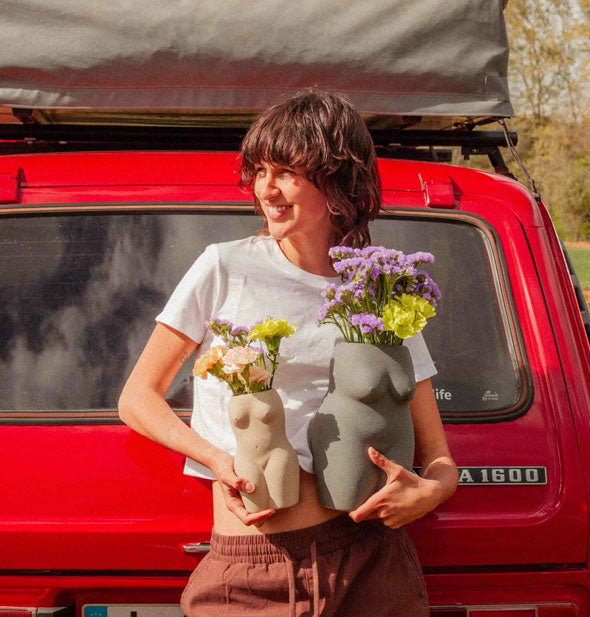 Smiling model standing in front of a red car holds a small beige nude form vase and a large gray nude form vase, both with fresh flowers inside